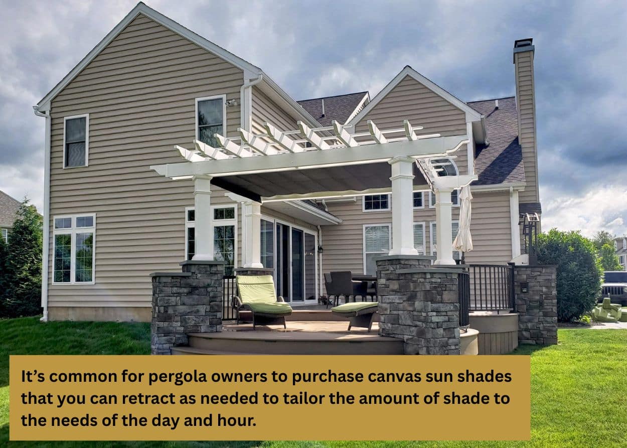 Backyard pergola attached to a home, built with white posts and stone pillars, providing shade over a raised deck. Caption: It’s common for pergola owners to purchase canvas sun shades that you can retract as needed to tailor the amount of shade to the needs of the day and hour.